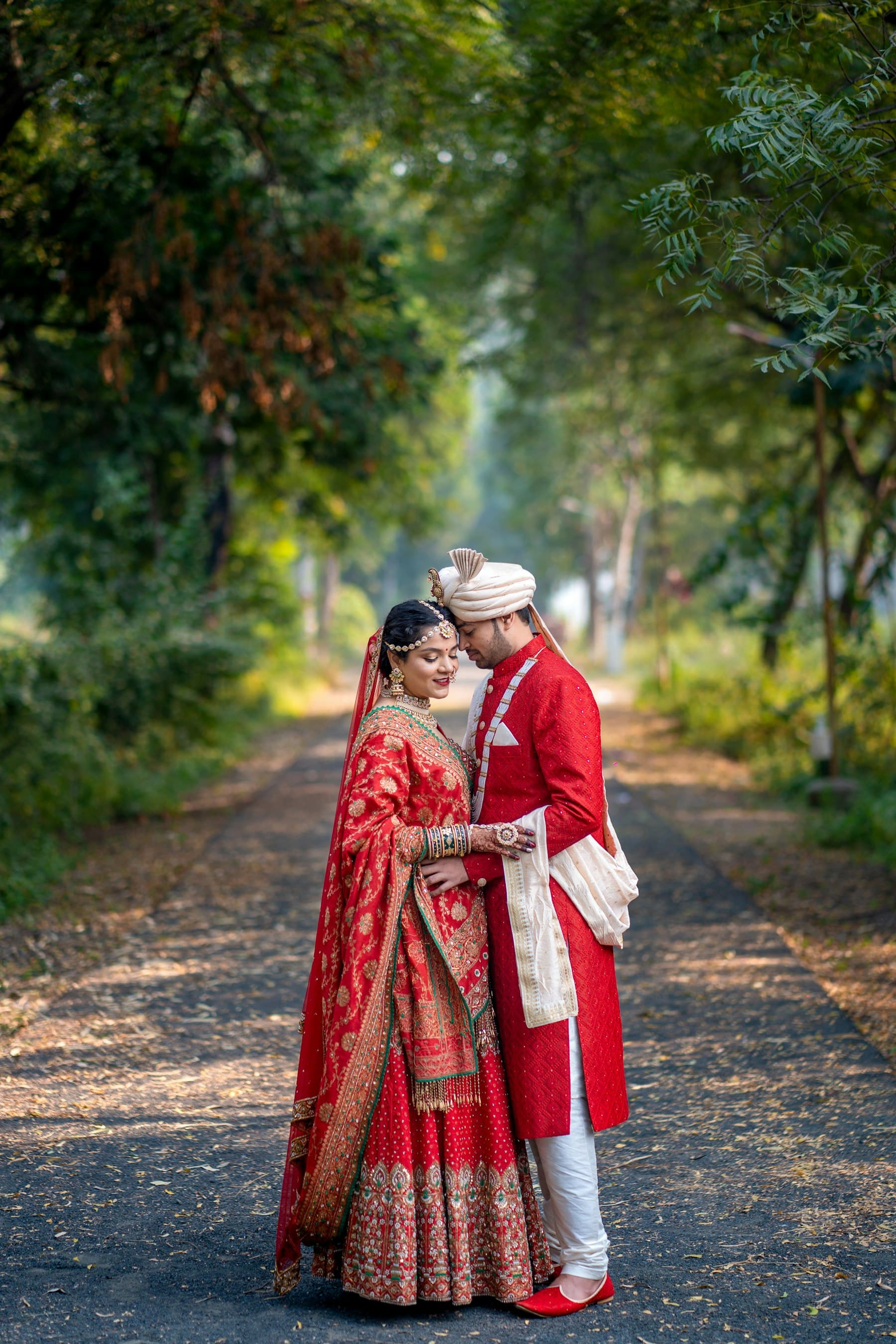 A couple in traditional red wedding attire walking together on a tree-lined path